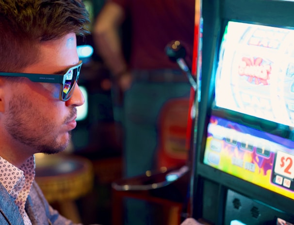 A man wearing sunglasses is intently focused on a slot machine, with colorful lights and graphics illuminating the screen. The scene is set indoors, likely in a casino or gaming area. His attire includes a patterned shirt and a blazer.
